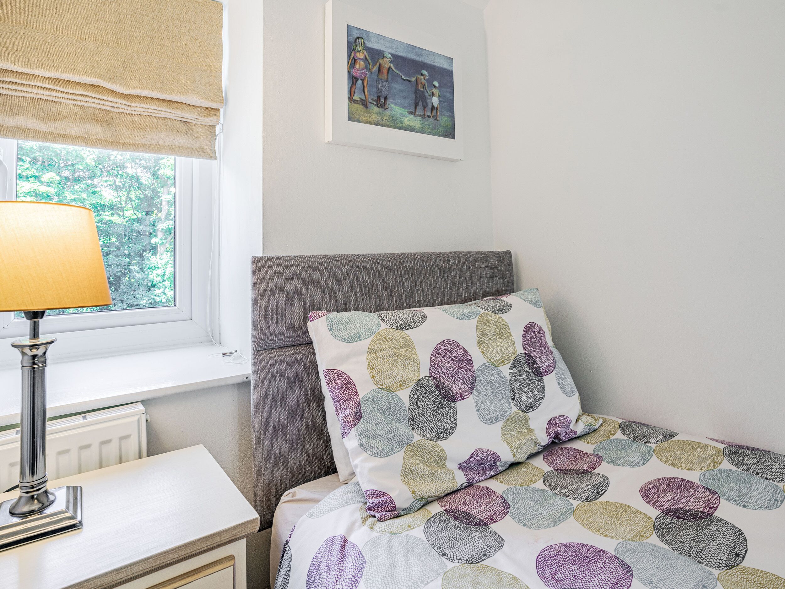 Een slaapkamer met een bed, nachtkastje, lamp, raam met uitzicht op bomen en een schilderij aan de muur.