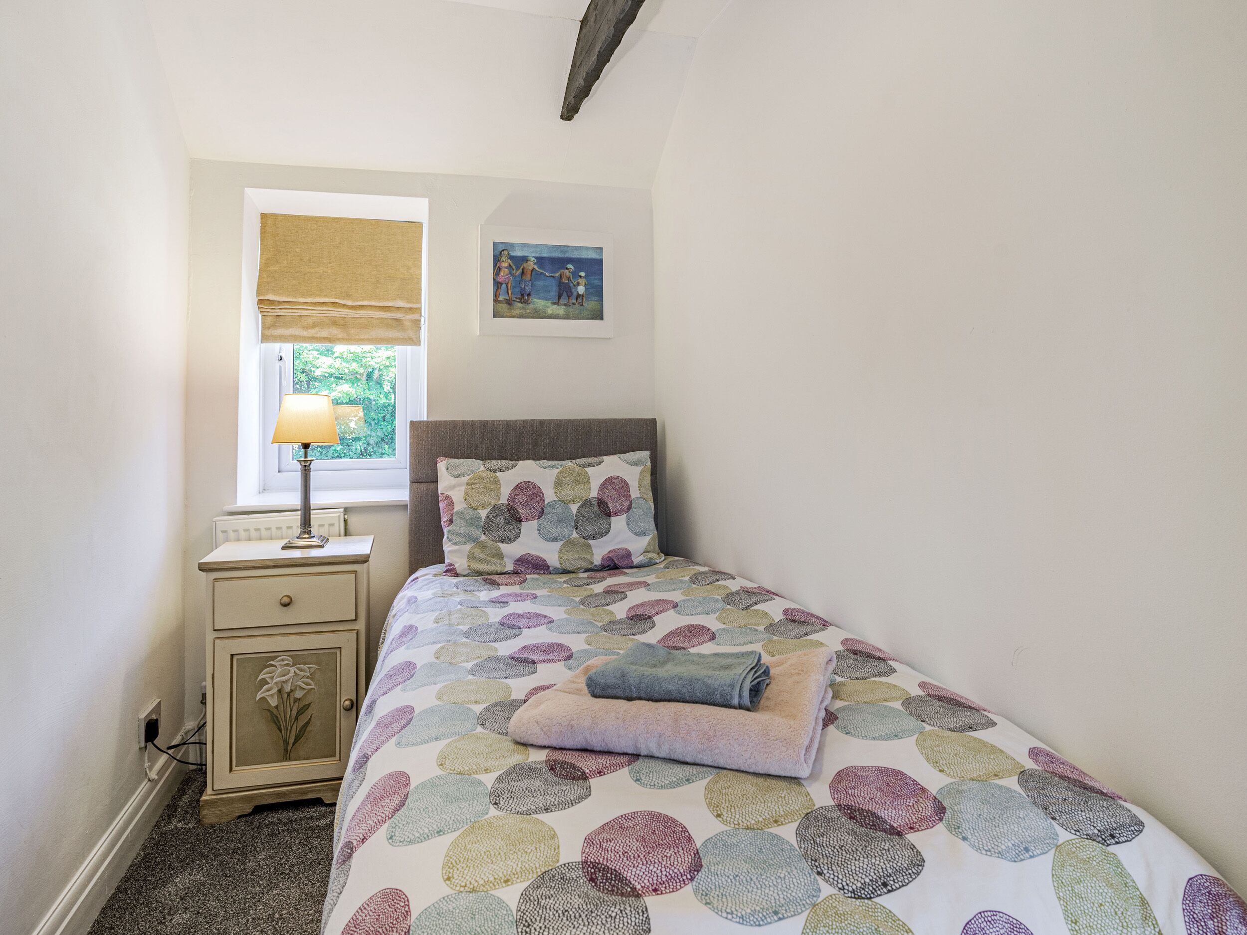 Un dormitorio con cama, mesita de noche, lámpara, ventana con vistas a zonas verdes y un cuadro en la pared.
