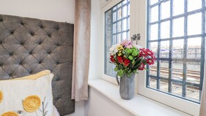 A bedroom with a tufted headboard, a window with a metal grid, and a vase of flowers on the windowsill. - George Stephenson @ Engine Shed (Whitby)
