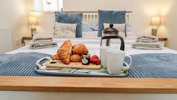 A neatly made bed with a wooden headboard, a bedside table with a lamp, and a tray with croissants, strawberries, and a coffee pot. - Bay Cottage (Filey)