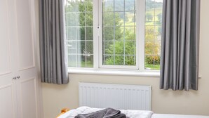 Quarto com cama, janela com cortinas, radiador e vista para o verde lá fora.