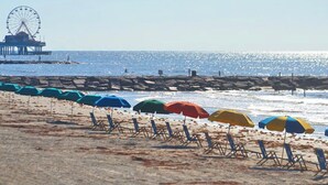 Una playa con hileras de sillas y sombrillas, un muelle con noria, y el océano al fondo.