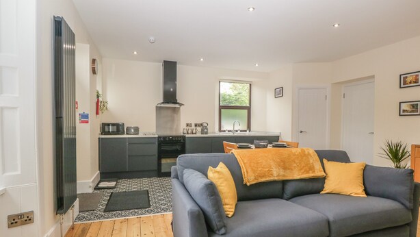 A modern living room with a grey sofa, wooden flooring, and a kitchen in the background. - Hillcrest (Newton Stewart)