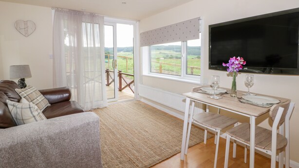 Una sala de estar acogedora con un sofá de cuero marrón, una mesa de comedor con dos sillas, un televisor montado en la pared y un ventanal con vistas al exterior.