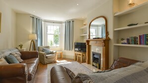 A living room with a fireplace, a television, a bookshelf, and a window with curtains. - Beachcomber (Padstow)
