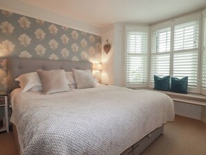 A bedroom with a large bed, a nightstand, a wall-mounted heart decoration, and a window with blinds. - Beachcomber (Padstow)