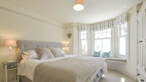 A neatly arranged bedroom with a large bed, bedside lamps, a window seat, and a view of the outside. - Beachcomber (Padstow)