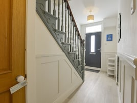 Un pasillo con una puerta de madera, una pared blanca, una puerta oscura, una lámpara y una estantería con un objeto azul.