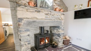 A stone fireplace with a wood-burning stove, a kitchen in the background, and a TV mounted on the wall. - Pen Y Braich Uchaf (Garndolbenmaen)