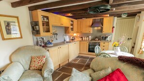 A cozy living room with a sofa and armchair, a dining table with chairs, and a kitchen area with wooden cabinets and appliances. - Old Stables Cottage (Saltburn-by-the-Sea)
