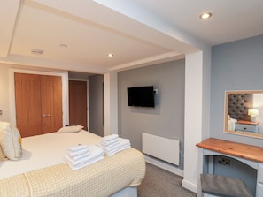 Quarto com cama, televisão na parede, mesa de cabeceira com luminária e porta de madeira.