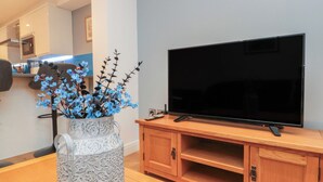 Un soggiorno moderno con un televisore a schermo piatto su un supporto in legno, un vaso decorativo con fiori blu e un angolo cucina sullo sfondo.