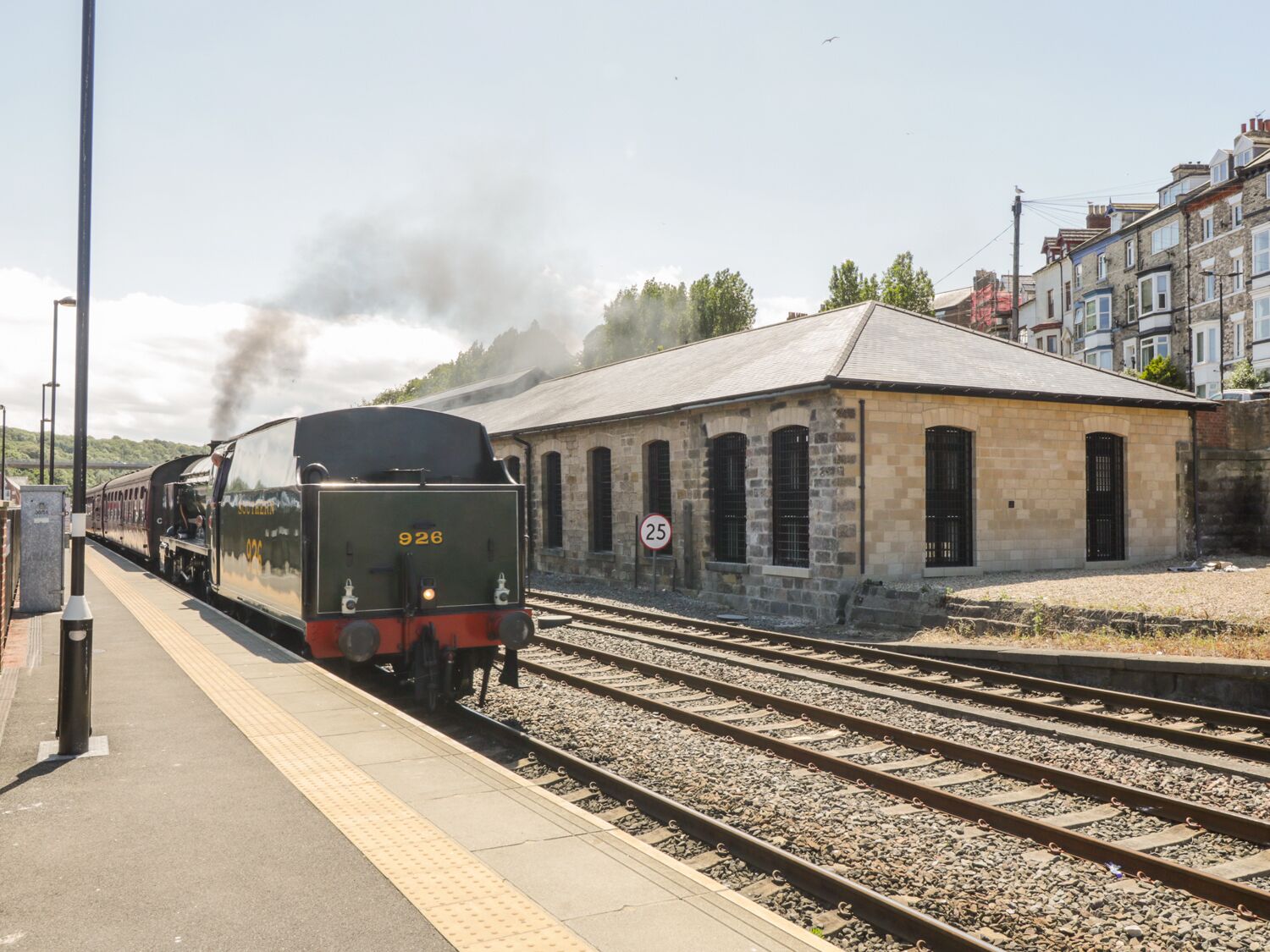 A steam train at a station with a stone building and multiple tracks.