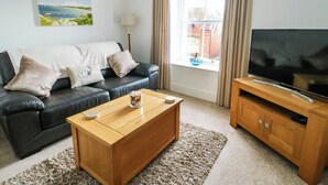Una sala de estar con un sofá de cuero negro, una mesa de centro de madera, un televisor sobre un mueble de madera y una ventana con cortinas.