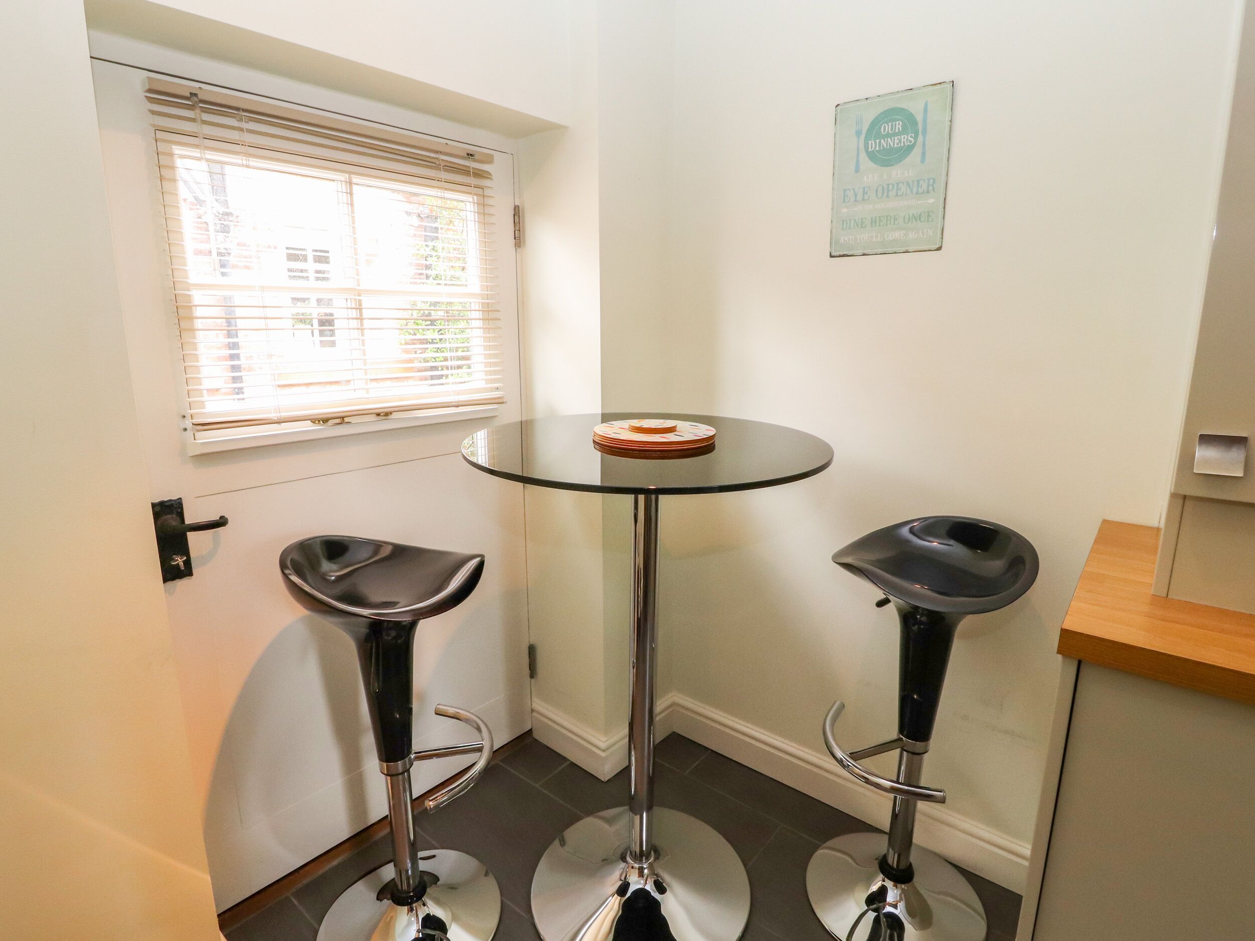A small room with a round glass table, two black bar stools, a window with blinds, and a sign on the wall.