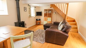A cozy living room with a wooden table, a brown leather sofa, a wooden staircase, and a black wood-burning stove. - The Dairy (Tarporley)
