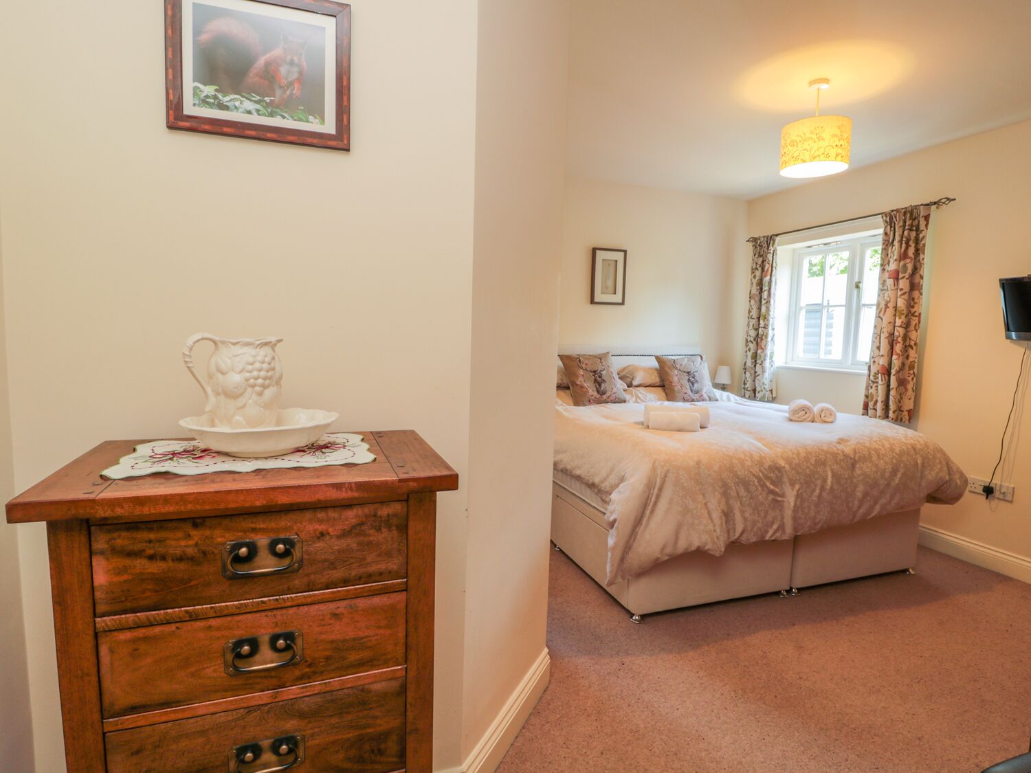 A bedroom with a bed, a dresser with drawers, a bedside table with a pitcher, a window with curtains, and a wall-mounted TV.