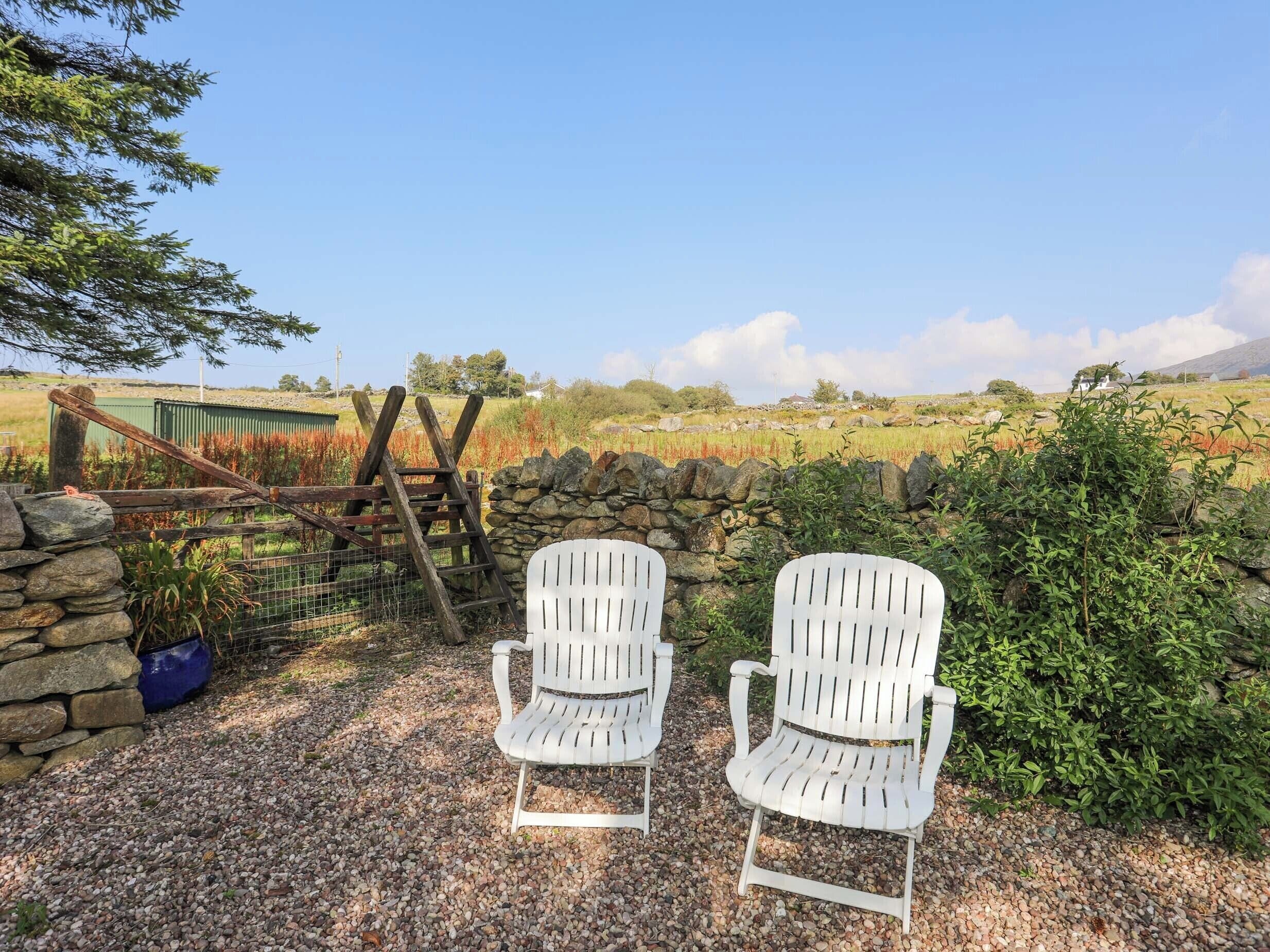 Two white Adirondack chairs on a gravel path with a stone wall and a wooden gate in the background.