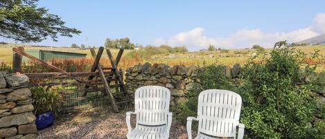 Two white Adirondack chairs on a gravel path with a stone wall and a wooden gate in the background.