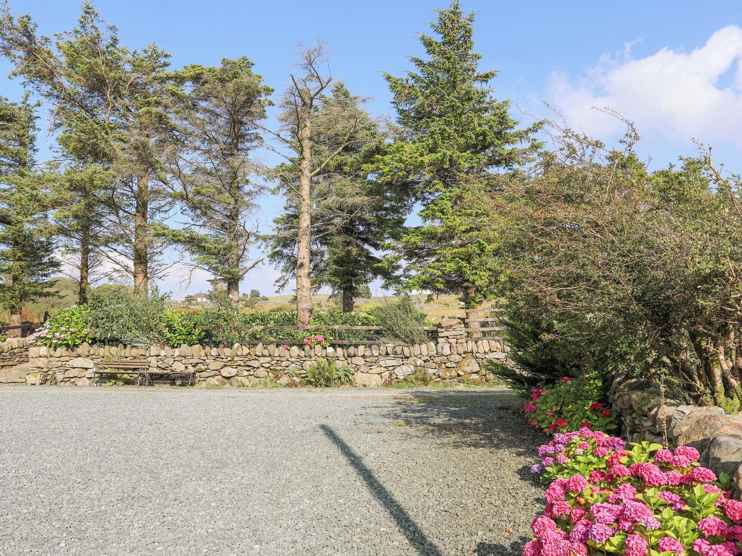 Une allée de gravier mène à un mur de pierres sur lequel est adossé un banc, le tout étant entouré de plantes en fleurs et de grands arbres.