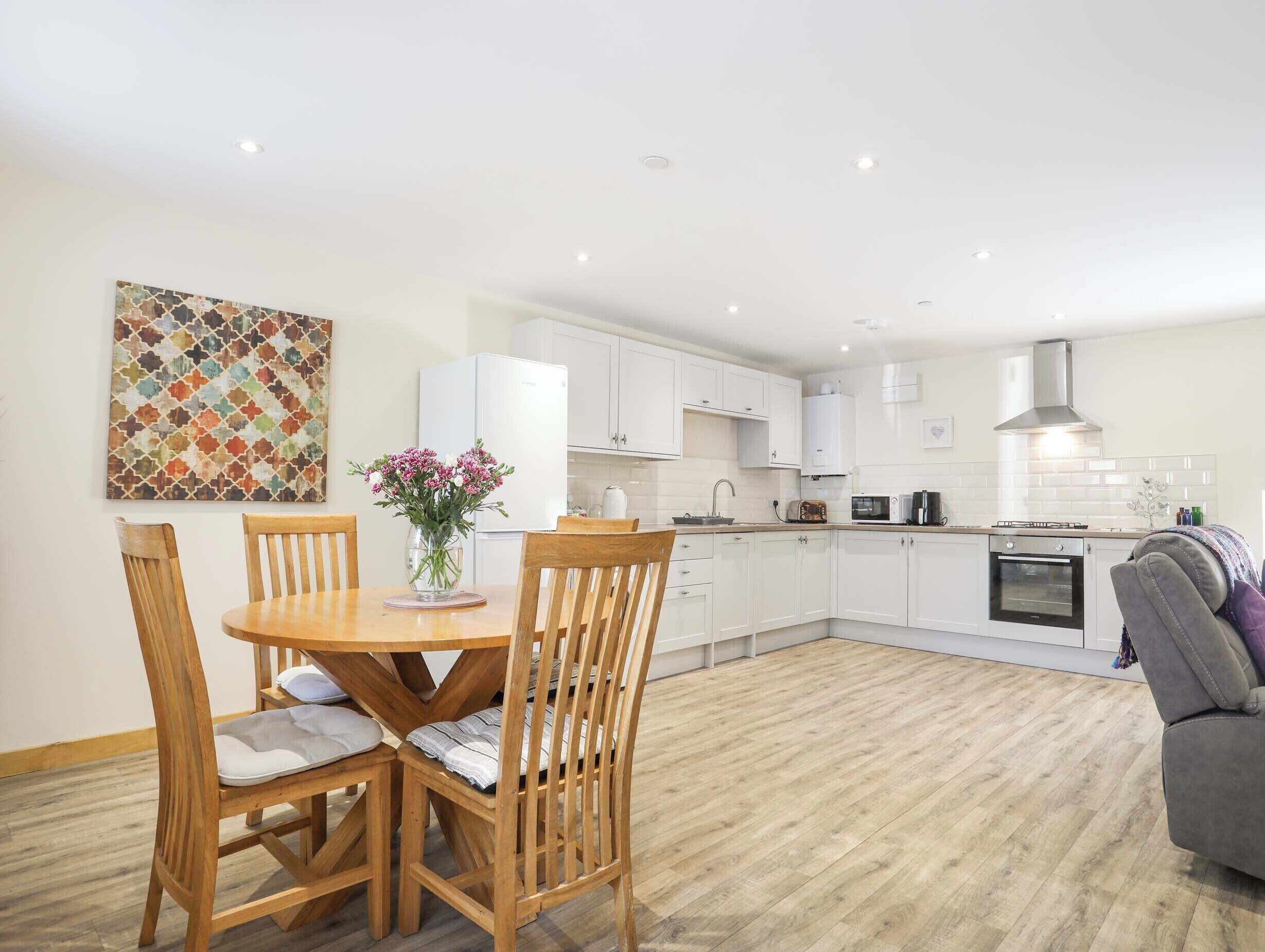 A modern kitchen with white cabinets, a built-in oven, and a microwave. There is a dining table with chairs and a vase of flowers.