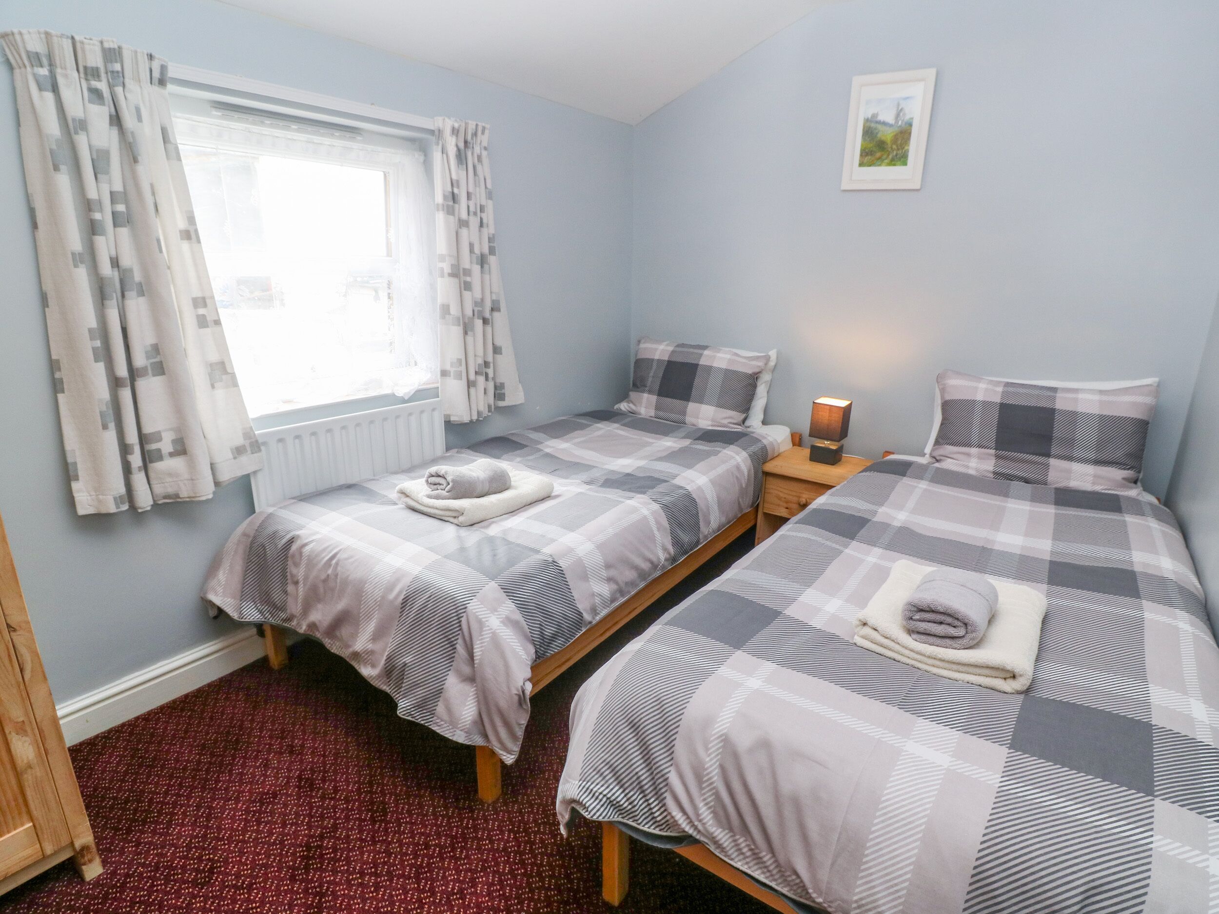 A bedroom with two beds, a window with curtains, a radiator, a bedside table with a lamp, and a framed picture on the wall.
