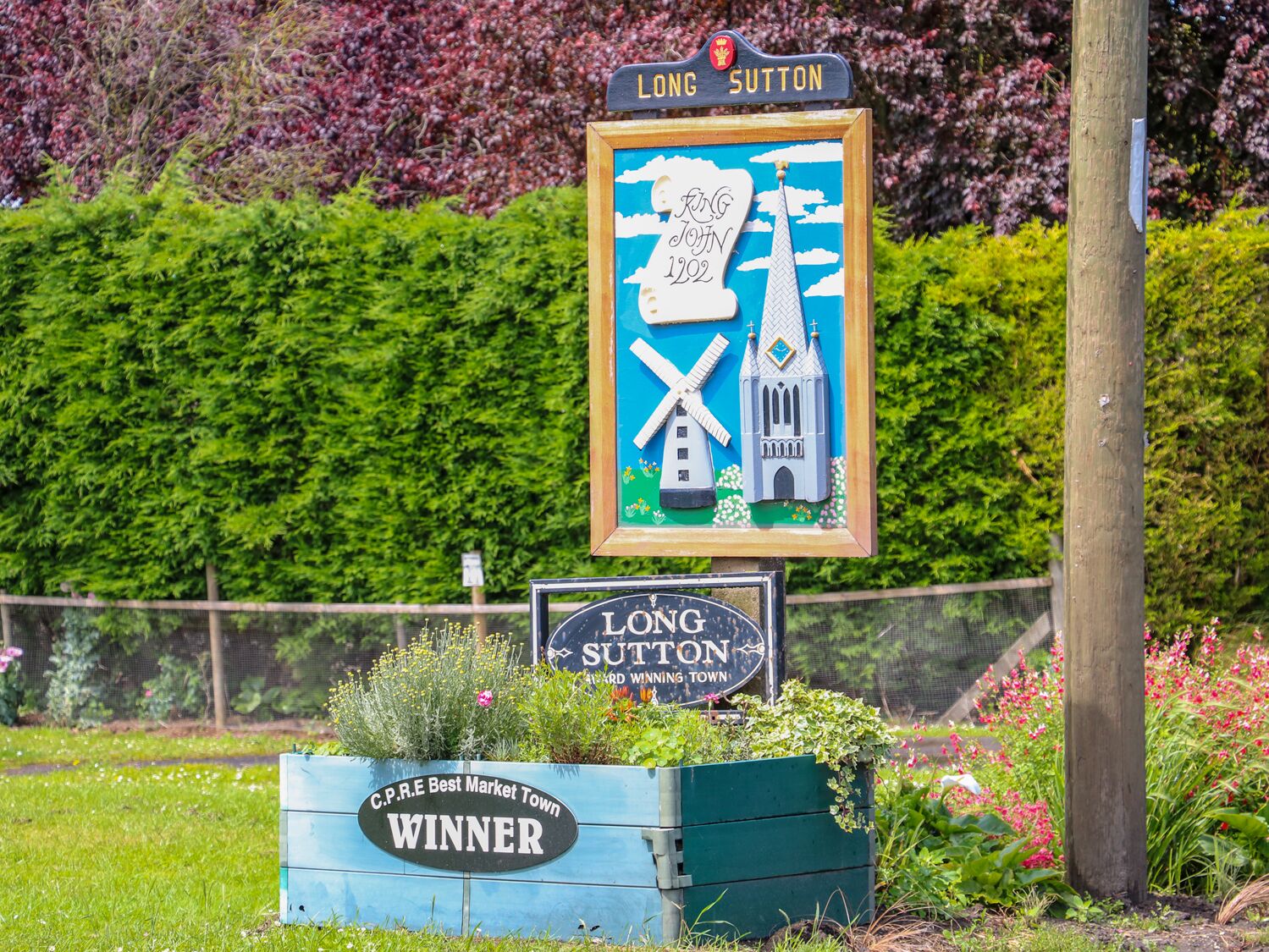 Ein Schild für Long Sutton, Gewinner des Wettbewerbs ‚Beste Marktstadt‘ des C.P.R.E, mit einer Darstellung einer Windmühle und einer Kirche.