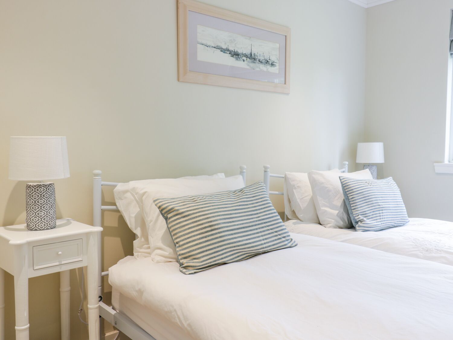 A bedroom with two beds, white bedding, striped pillows, a bedside table with a lamp, and a framed picture on the wall.