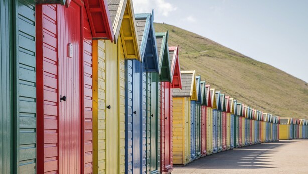 A row of colorful beach huts with a grassy hill in the background.
