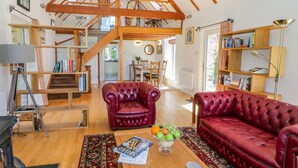 A spacious living room with a high ceiling, wooden beams, a staircase, leather sofas, a coffee table with magazines, and a bookshelf. - The Barn at Waen y Ffynnon (Ruthin)