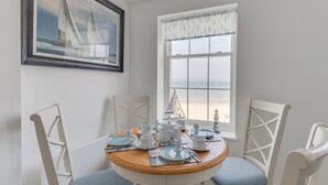Un coin repas avec une table ronde dressée pour quatre personnes, des chaises blanches, un tableau représentant un voilier et une fenêtre donnant sur la plage.
