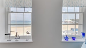 Deux fenêtres donnant sur la plage, chacune agrémentée d’un rideau et d’une petite étagère sur laquelle sont disposés des objets décoratifs.