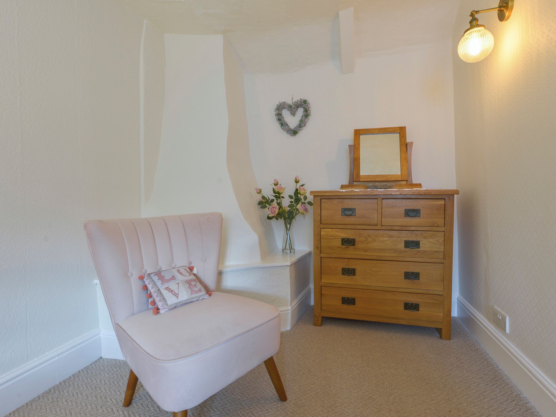 A room with a pink chair, a wooden dresser with drawers, a small table with a vase of flowers, and a wall-mounted light fixture.