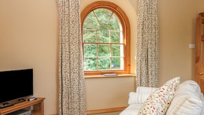 A living room with a television on a wooden stand, a sofa with patterned cushions, and a window with curtains. - Elmwood (Buxton)