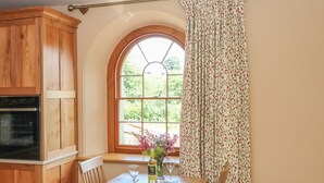 A kitchen with wooden cabinets, a built-in oven, a dining table with chairs, and a window with a view of greenery. - Elmwood (Buxton)