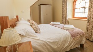 A neatly made bed with pillows and a blanket, a bedside lamp, and a window with curtains. - Elmwood (Buxton)