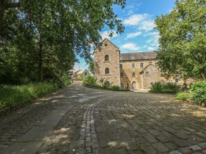 A historic stone building with a central tower, surrounded by lush greenery and a cobblestone path.