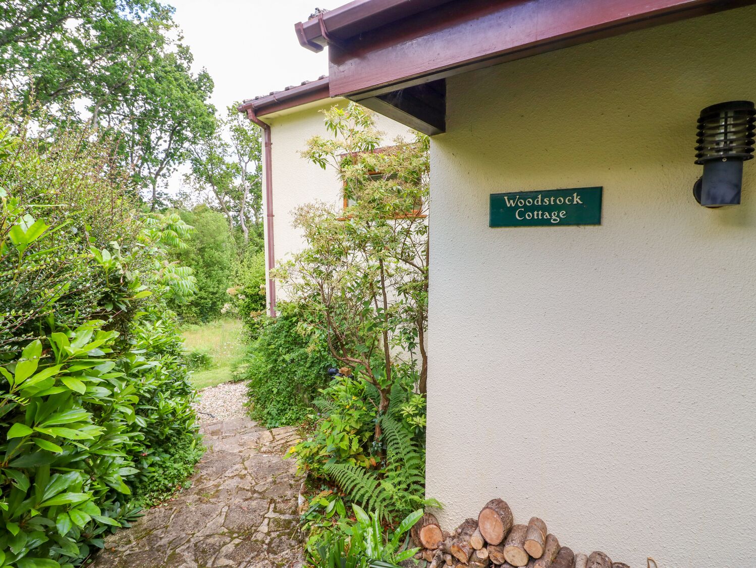 Un chemin menant à un bâtiment portant l’inscription « Woodstock Cottage », entouré d’une végétation luxuriante et d’un mur de pierre.