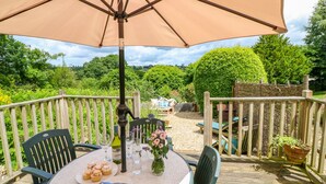 Un espace de restauration en plein air avec une table ronde, des chaises, un grand parasol et une vue sur un jardin doté d’une terrasse en bois et de verdure.