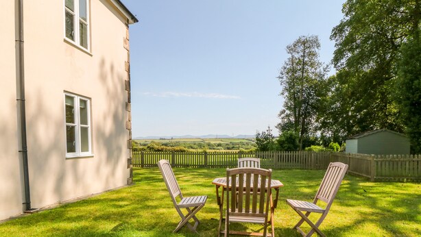 A patio with a table and four chairs, a stone building, and a view of a green landscape. - Tirionfa (Llangefni)
