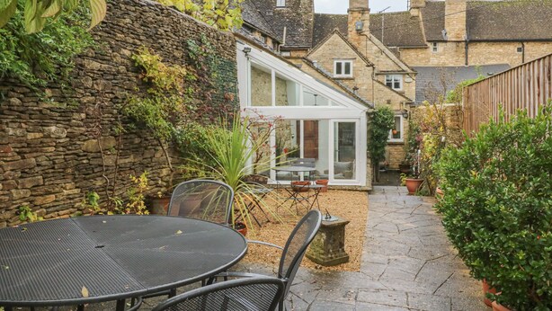 Un mur de pierres recouvert de lierre, une terrasse avec une table et des chaises, et une véranda offrant une vue sur les maisons voisines.