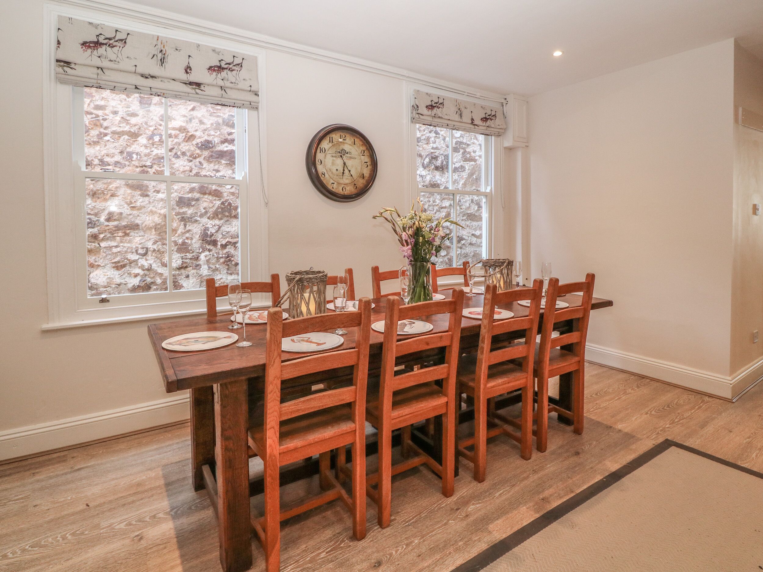 A dining room with a wooden table set for six, flanked by two windows with decorative valances, a wall clock, and a vase with flowers.