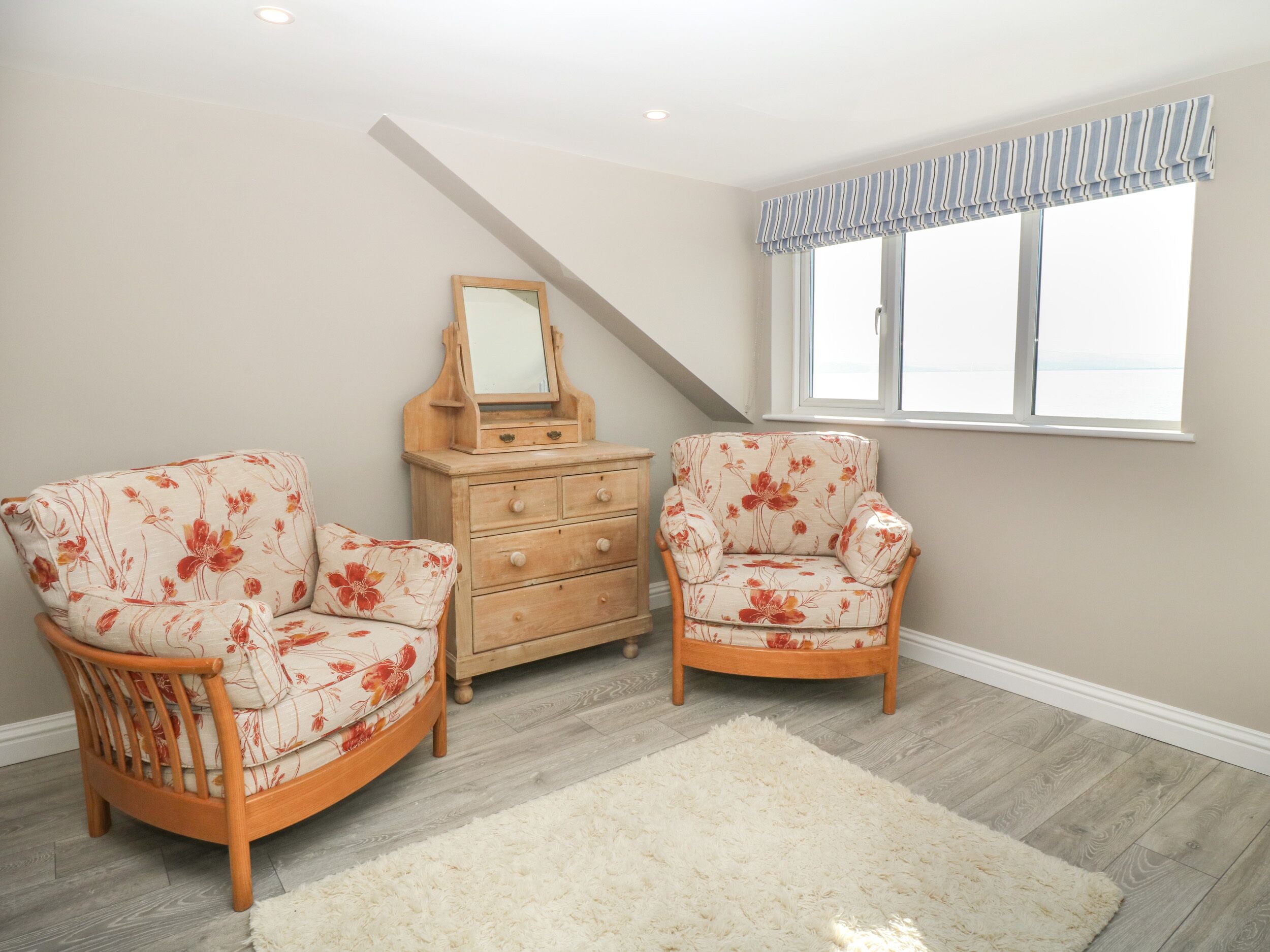 Een kamer met twee bloemenarmstoelen, een houten dressoir met spiegel en een groot raam met gestreept gordijn.