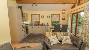 Una sala de estar acogedora con un sofá de cuero marrón y un sillón gris, un comedor con mesa y sillas, una cocina con armarios blancos y un refrigerador, y un ventanal con cortinas.