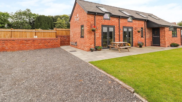A brick building with a sloped roof, a patio with a table and chairs, and a fenced area. - The Shippon (Tarporley)