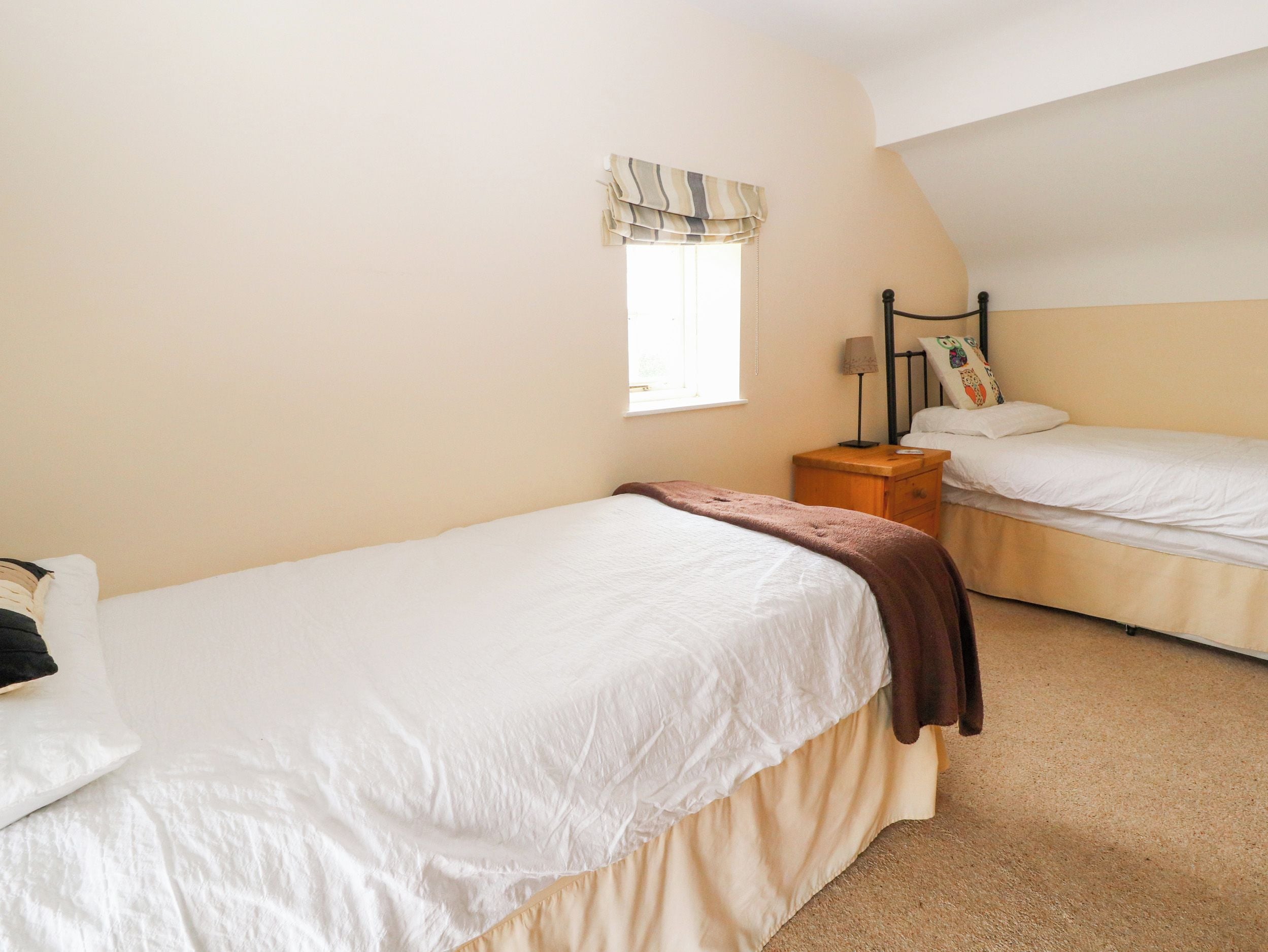 Un dormitorio con dos camas individuales, una ventana con cortina a rayas, una mesita de noche con lámpara y una estantería en la pared.