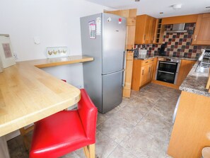 A compact kitchen with a refrigerator, wooden cabinets, a tiled backsplash, and a wooden dining table with a red chair. - The Shippon (Tarporley)