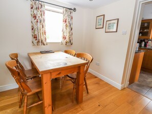 A dining area with a wooden table and chairs, a window with floral curtains, and a kitchen visible in the background. - The Shippon (Tarporley)