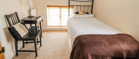 A small, well-lit room with a single bed, a desk with a chair, and a window with blinds.