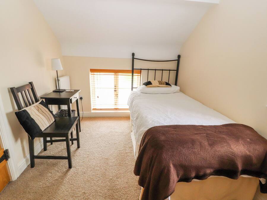 A small, well-lit room with a single bed, a desk with a chair, and a window with blinds.